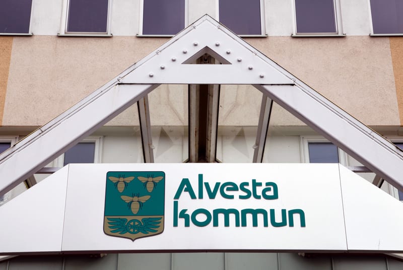 En skylt med texten "Alvesta kommun" och kommunens vapen, som föreställer tre bin och en bevingad hjul, är monterad under ett vitt tak. Bakom skylten syns en byggnadsfasad med fönster.