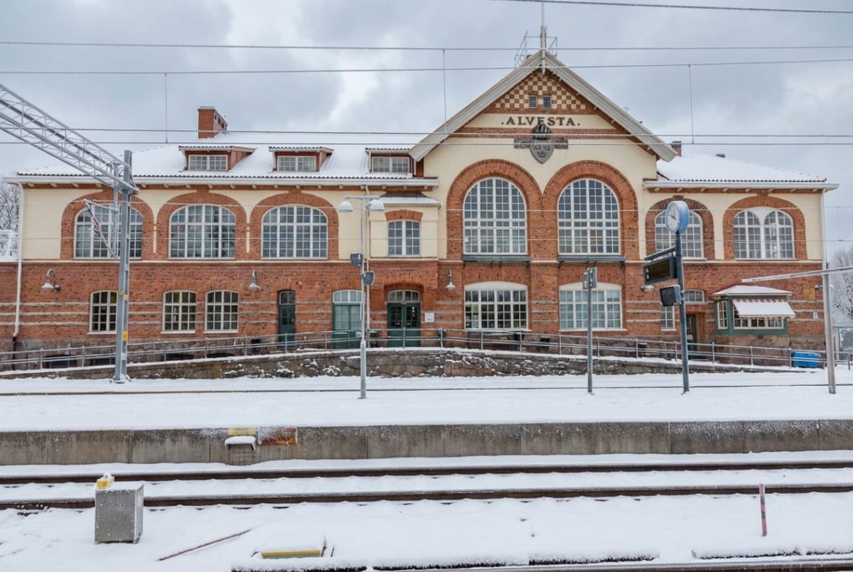 En bild av en tegelbyggnad med vita detaljer och skylten "ALVESTA" på fasaden, tagen från ett tågspår som är täckt av snö. Byggnaden har många stora fönster med välvda överdelar och taket är också snötäckt.