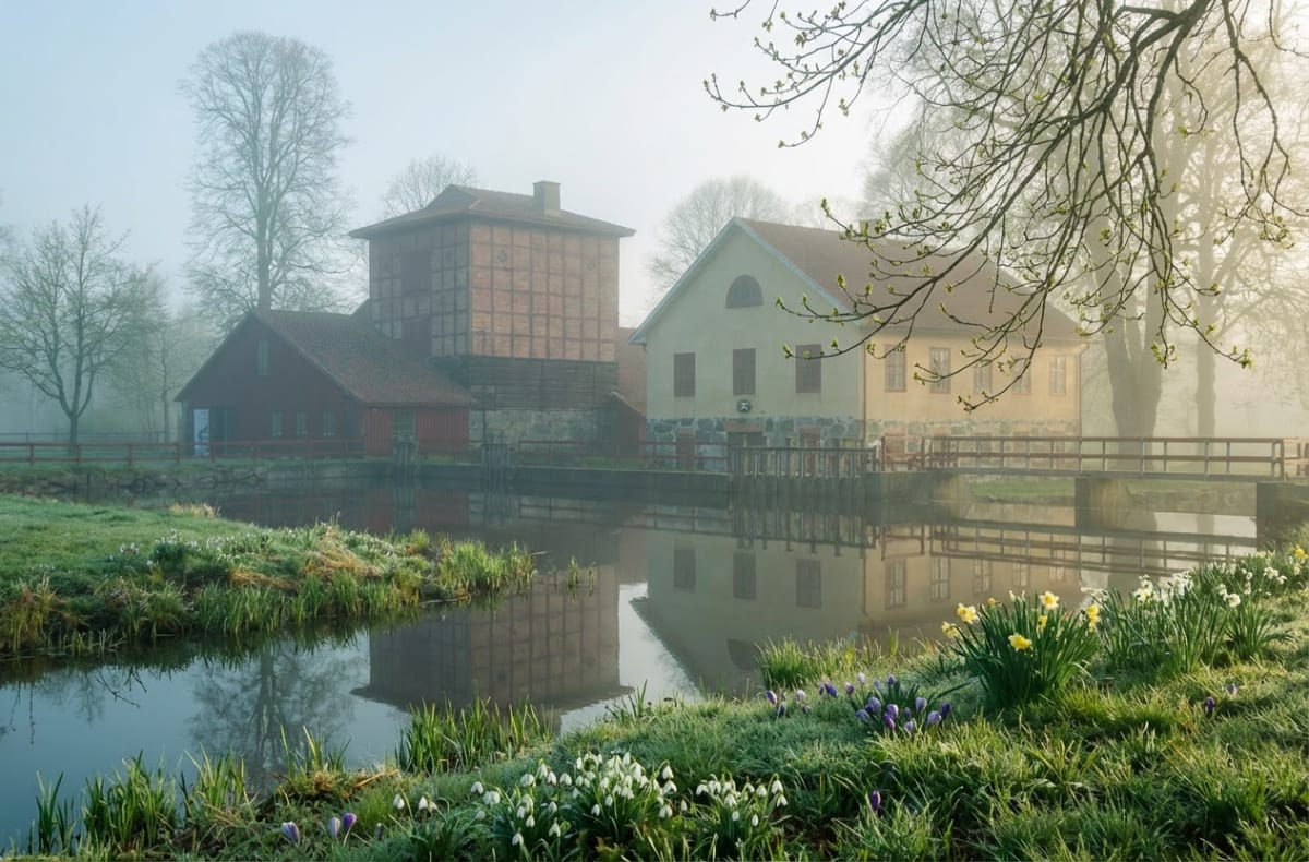 En bild en dimmig vårdag föreställande ett vattendrag med byggnader och grönska. Vattendraget reflekterar flera byggnader med röda tak och fasader i rött tegel samt ljusgul puts. På stränderna syns gräs, snödroppar, krokusar och påskliljor.