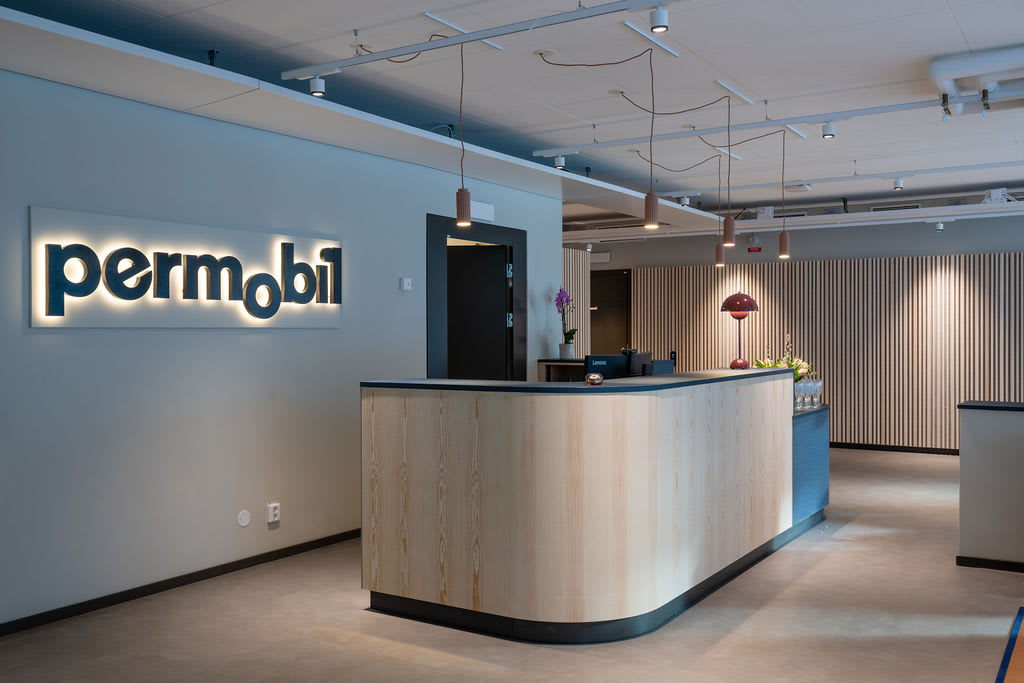 En bild interiört som visar en reception med en stor, upplyst Permobil-logotyp på väggen. Till höger finns en träfärgad receptionsdisk med en röd lampa, en dörröppning och en blomsterarrangemang.