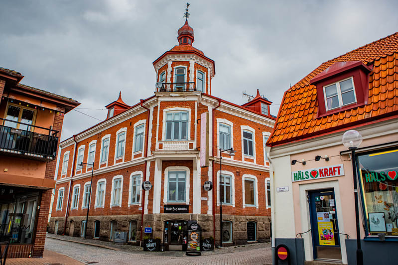 En bild som visar en stadsmiljö med flera byggnader i olika färger. I förgrunden till höger finns en ljusrosa byggnad med texten "HÄLS❤KRAFT" på en skylt. I mitten syns en röd tegelbyggnad med vita fönsterkarmar och en röd kupol på taket. En butik me