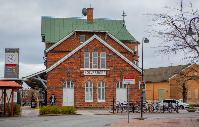 En stor, röd tegelbyggnad med ett grönt tak dominerar bilden. Byggnaden har vita fönster och dörrar samt skylten "SÖLVESBORG" på fasaden. Till vänster om byggnaden finns en klocka och informationstavla, och framför den står flera cyklar parkerade. En