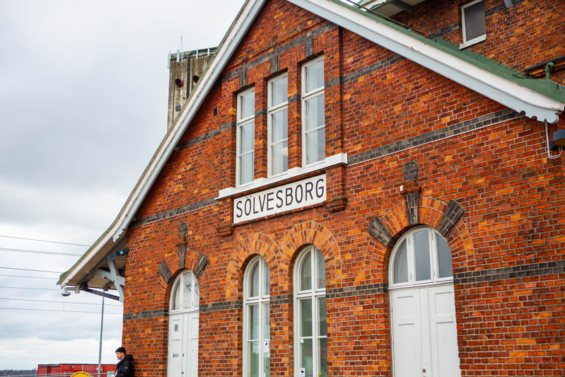 En tegelbyggnad med vita fönster och dörrar har en skylt med texten "SÖLVESBORG" synlig. En person står till vänster i bild framför byggnaden under en mulen himmel. Byggnaden har ett utkragande tak och tegelväggarna är i varierande röda och mörkare n
