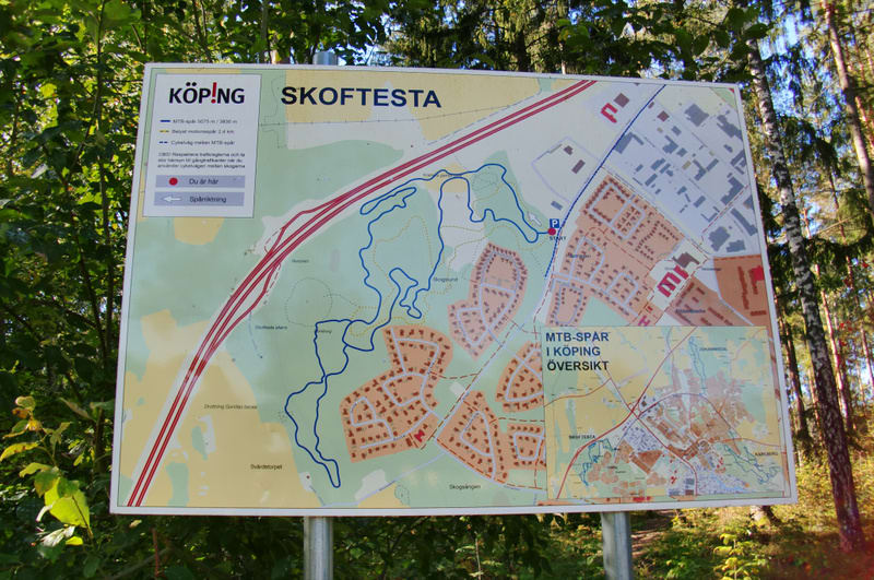 En stor skylt med en karta över Köping och MTB-spår, omgiven av grönska och träd. Kartan visar terräng med bostadsområden, skogar, samt information om MTB-leder och en "Du är här"-markör. Skogen syns i bakgrunden.