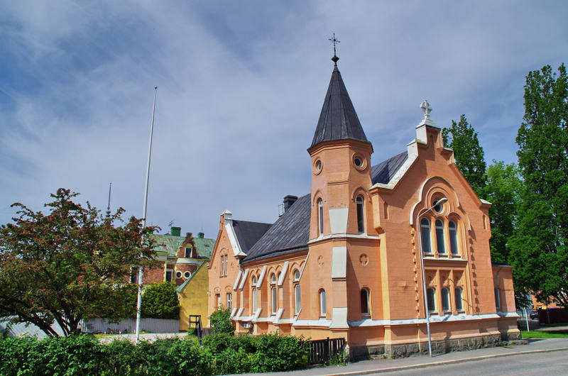 En stor, persikofärgad byggnad med svart tak och torn i gotisk stil står intill en väg med en gräsbevuxen trottoarkant. Bakom byggnaden syns träd och andra hus. Himlen är blå med vita moln och en flaggstång skymtar till vänster.
