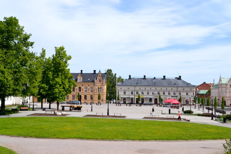 En översiktsbild av ett torg med gräsmattor och träd i förgrunden, omgivet av byggnader i bakgrunden. Flera personer, parkerade bilar och en röd paviljong syns på torget.