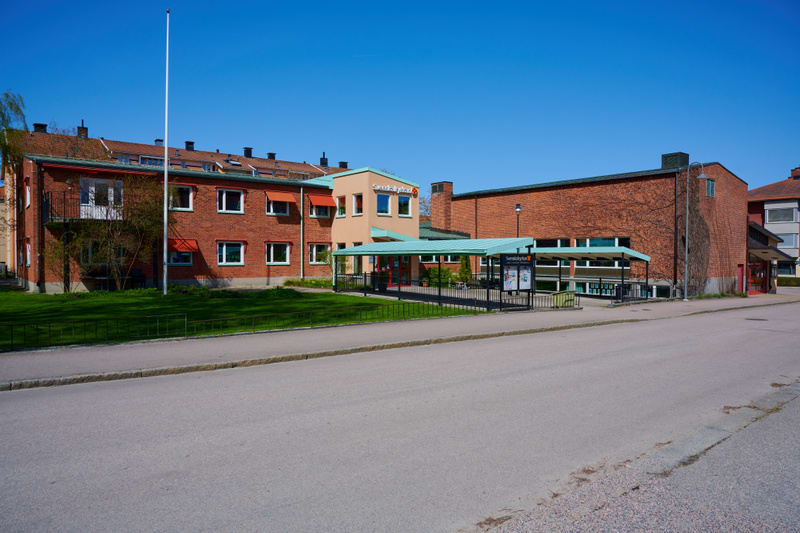 En fasadvy av en tegelbyggnad med flera fönster och röda markiser, bredvid en ljusare byggnadsdel med grönt tak. Framför byggnaden finns ett grönområde, en flaggstång, en väginformationstavla och en asfalterad väg. Himmelen är klarblå.