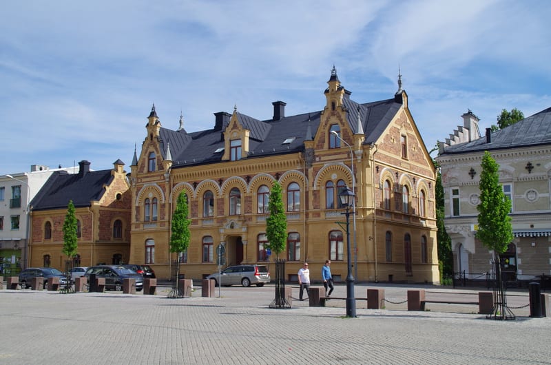 En bild av en torgplats med ett stort ockrafärgat hus med mörkt tak. Framför huset parkerade bilar och två personer som promenerar. Flera unga träd kantar torgplatsen. En gatlykta står till höger i bilden och himlen är blå med sprida moln.