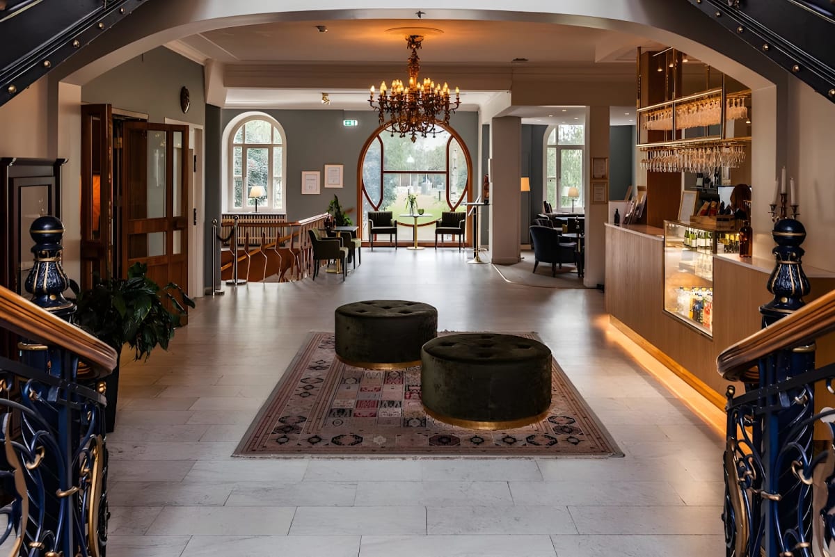 Bilden visar ett inomhusmiljö med en foajé eller hotellobby, sett från nedre delen av en trappa. I förgrunden syns ett mönstrat golv och en matta med två cylinderformade fotpallar i mörk färg. En stor kristallkrona hänger i taket, och framför den fin