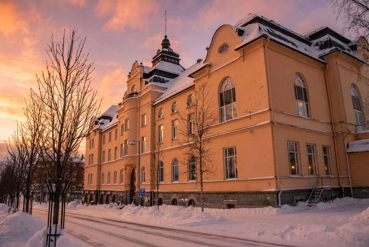 En gul byggnad med många fönster och snö på taket syns under en himmel med rosa och orangea moln. Framför byggnaden finns en snötäckt gata med spår samt flera trädstammar med bara grenar.