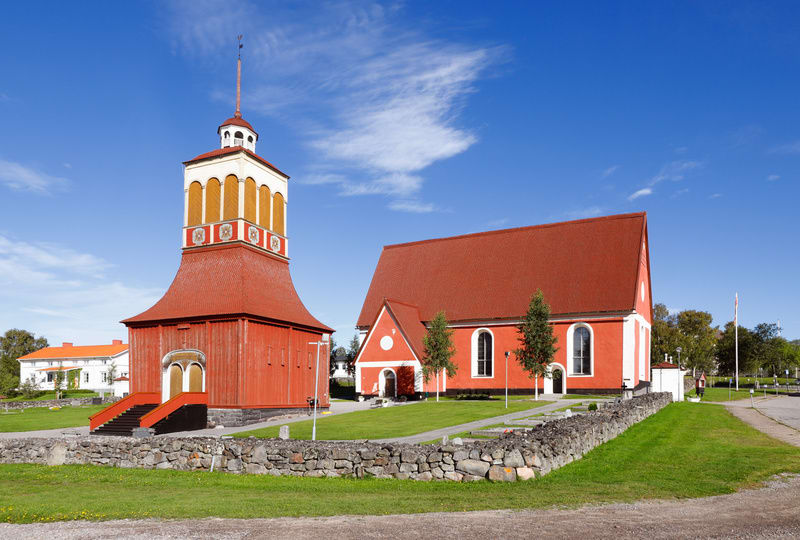 En röd kyrkobyggnad och en matchande klockstapel står på en grön gräsmatta under en klarblå himmel. Stenmurar och grusvägar omger området, och det finns träd och mindre byggnader i bakgrunden. Klockstapeln har en gul och vit del överst med ett spetsi