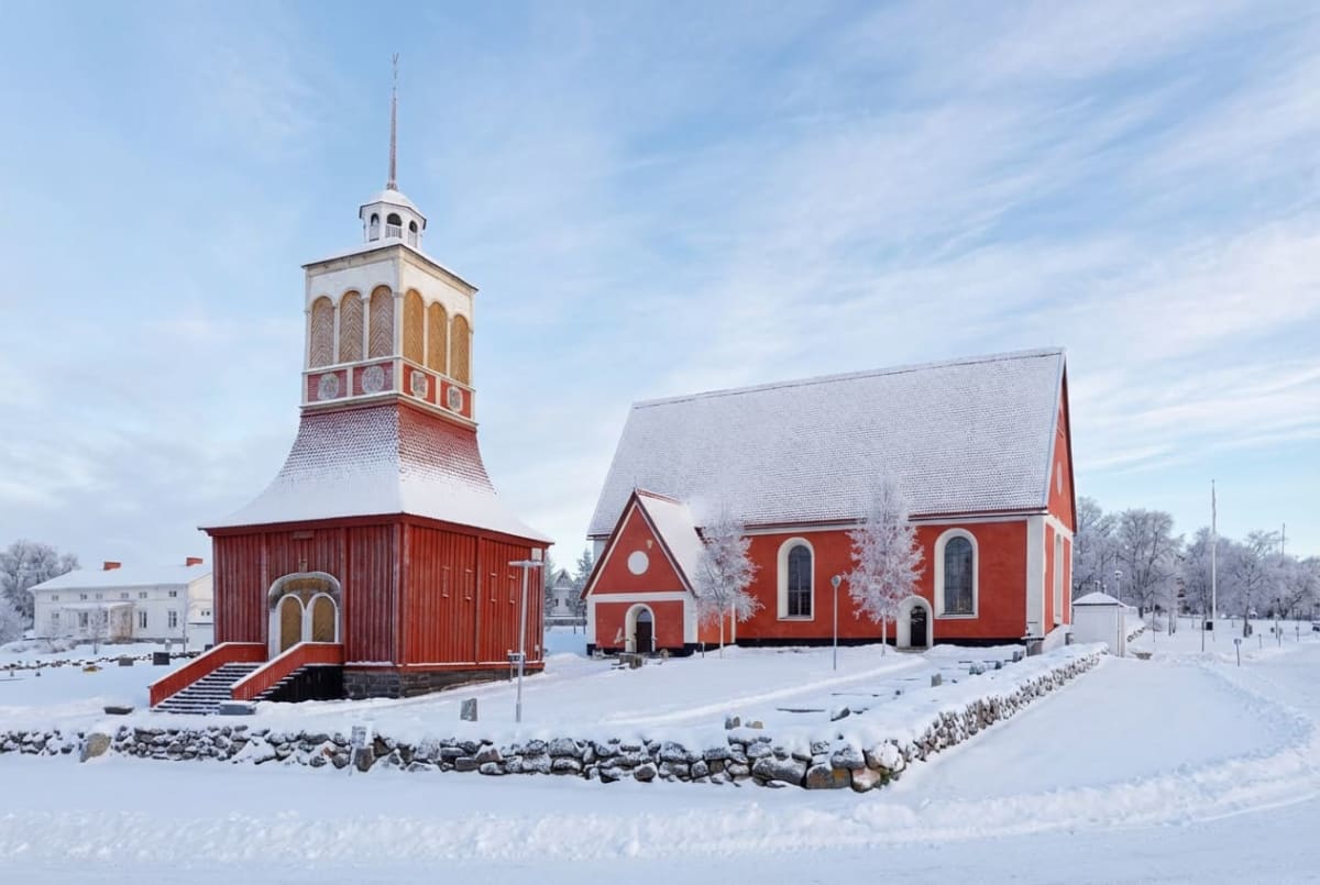 En röd träkyrka med ett fristående klocktorn står i ett snötäckt landskap under en klarblå himmel. Tak och mark är täckta av snö, och det finns ett staket av stenar i förgrunden samt snötäckta träd i bakgrunden.