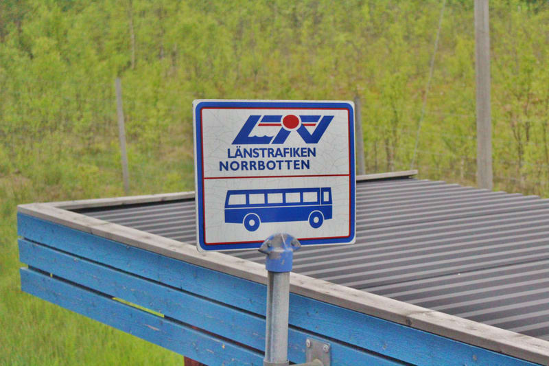 En busskylt för Länstrafiken Norrbotten står vid en blå busskur med ett tak av korrugerad plåt. I bakgrunden syns grönskande träd och buskar, vilket indikerar att bilden är tagen utomhus under en varmare årstid.