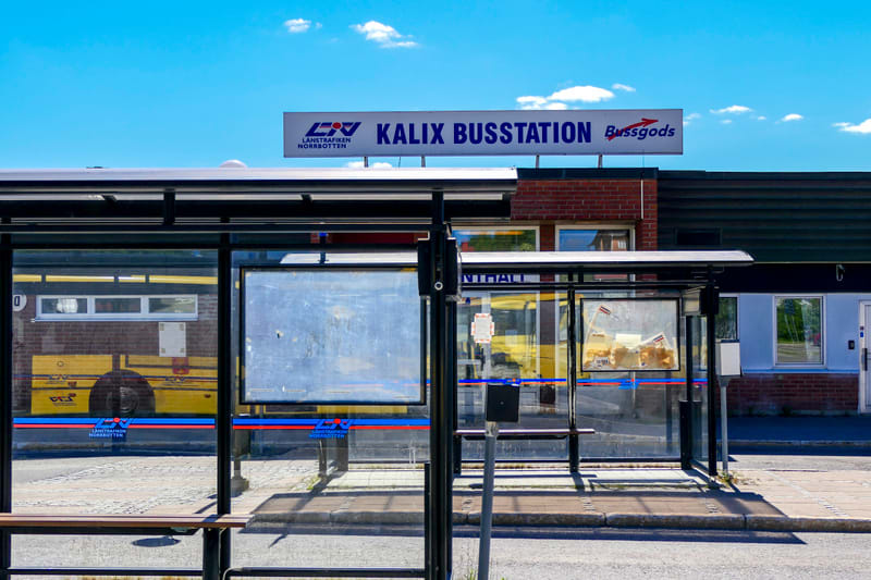 En bild som visar en busshållplats och en busskur i förgrunden, med en byggnad och en skylt med texten "Kalix Busstation" och "Länstrafiken Norrbotten" i bakgrunden. En gul buss syns reflekterad i busskurens glas.
