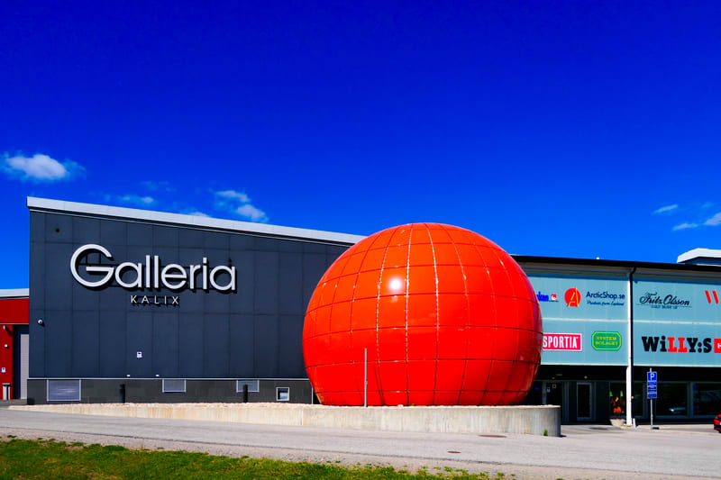 En modern byggnad med texten "Galleria Kalix" på en mörkblå fasad. Framför byggnaden står en stor, röd, sfärisk konstruktion. Till höger syns en annan del av byggnaden med flera skyltar för butiker. Hela scenen är under en klarblå himmel med några vi