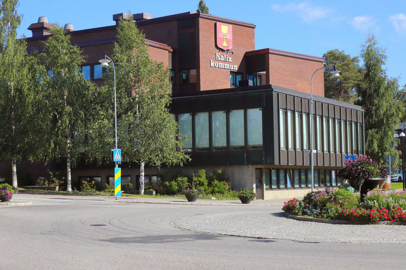 En stor byggnad med tegelfasad, troligtvis kommunhus, skyltad med "Kalix kommun" och kommunvapen. Framför byggnaden finns träd, en cirkulationsplats med blomsterplanteringar, en lyktstolpe och en övergångsskylt.