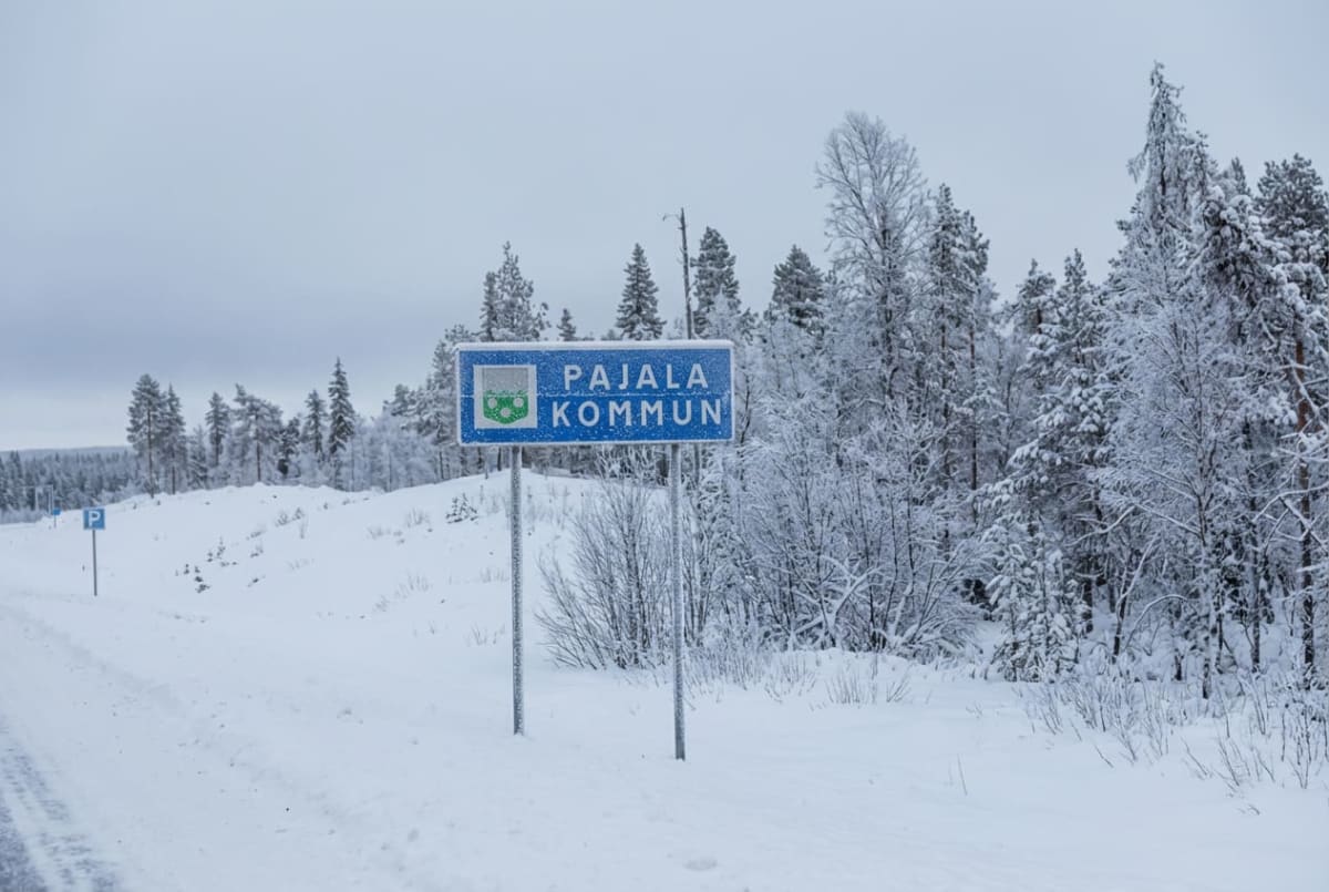 En blå och grön skylt med texten "PAJALA KOMMUN" och ett kommunvapen står i förgrunden vid en snötäckt väg. I bakgrunden syns snöklädda träd under en molnig himmel. Marken är täckt av snö.