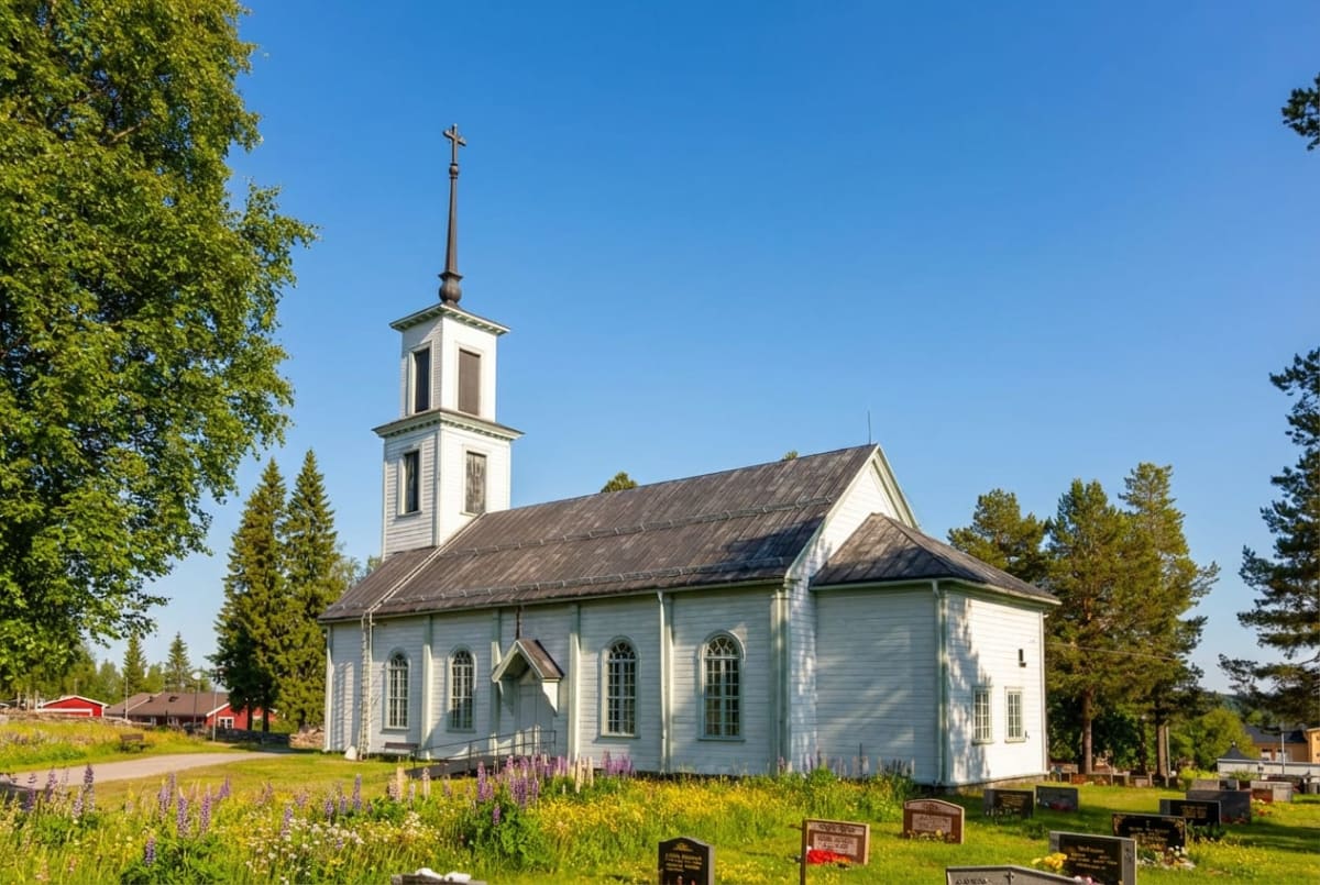 En vit kyrkobyggnad med ett klocktorn och en spira med ett kors ovanpå, omgiven av grönska, blommor och några gravstenar. Bakom kyrkan syns träd och i bakgrunden skymtar röda byggnader under en klarblå himmel.