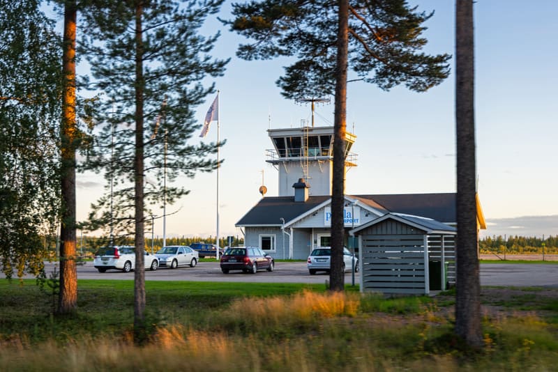 En bild av en flygplatsbyggnad med ett kontrolltorn, omgiven av träd och parkerade bilar. Ett flertal flaggstänger med flaggor syns bredvid byggnaden. Marken består av gräs och grus. Himlen är ljus med några moln.