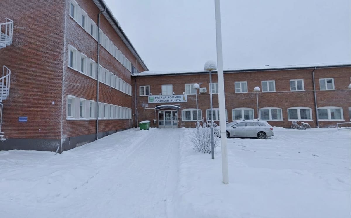 En fasadbild av en större tegelbyggnad under vintertid. Byggnaden har många fönster och en skylt ovanför huvudentrén där det står "PAJALA KOMMUN PAJALAN KUNTA". Det ligger snö på marken, på ett fordon och på växtligheter.
