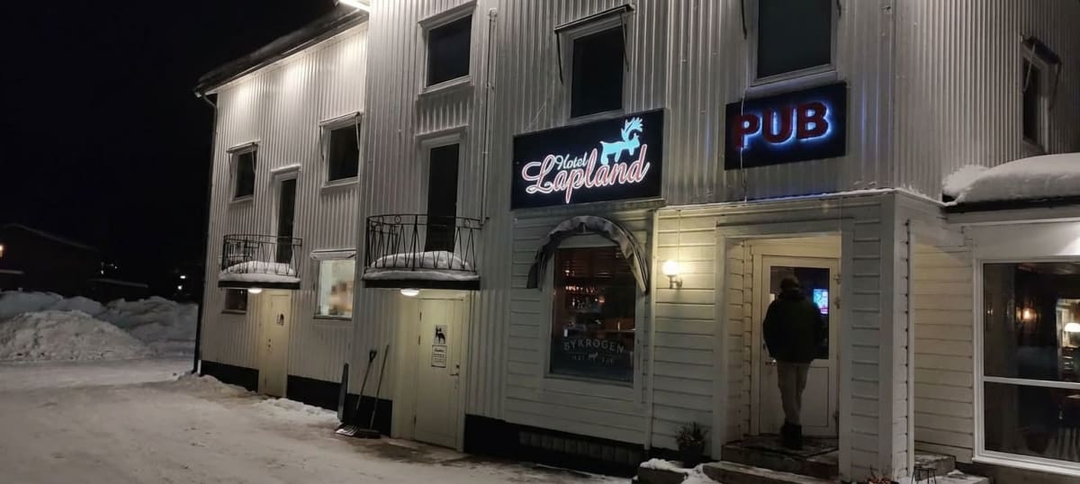 En vit träbyggnad med flera fönster och dörrar under en mörk vinterkväll. Skyltar med texten "Hotell Lapland" och "PUB" är upplysta. Vid en dörr står en person, och snöiga drivor syns runt byggnaden.