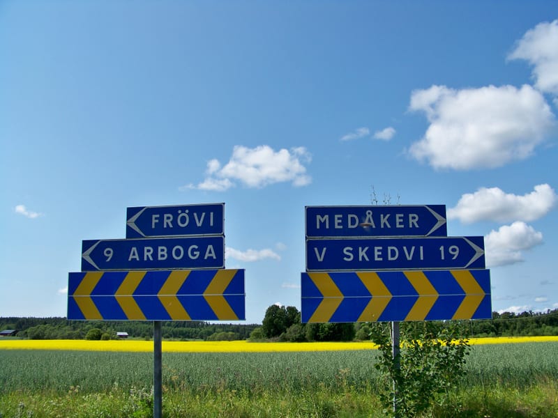 Två blåa vägskyltar med vita text och pilar står framför stora fält. Den vänstra skylten visar vägen mot "FRÖVI" och "9 ARBOGA". Den högra skylten pekar mot "MEDÅKER" och "V SKEDVI 19". Fälten består av grön gewäsxt och lysande gul raps. I bakgrunden