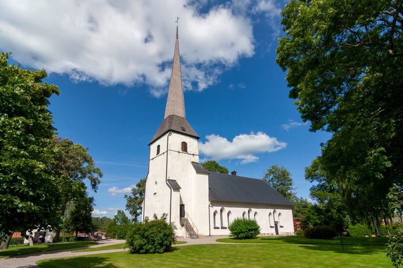 En vit kyrkobyggnad med en hög, spetsig spira står under en klarblå himmel med vita moln. Grön gräsmatta framför kyrkan och lummiga träd ramar in bilden. En grusgång leder förbi byggnaden.