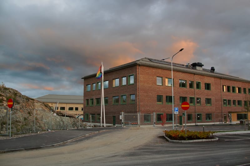 En brickbyggnad med gröna fönsterramar under en molnig himmel med orangea inslag, bredvid en stenig kulle. En regnbågsflagga är hissad på en flaggstång framför byggnaden. Parkeringsplatser och trafikskyltar syns också.