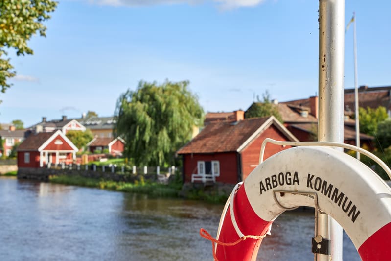 En bild som visar en vit och röd livboj med texten "ARBOGA KOMMUN" i förgrunden, fastsatt vid en metallstolpe. I bakgrunden syns ett vattendrag med flera röda stugor längs stranden under en klarblå himmel med några moln. Träd och grönska syns också.