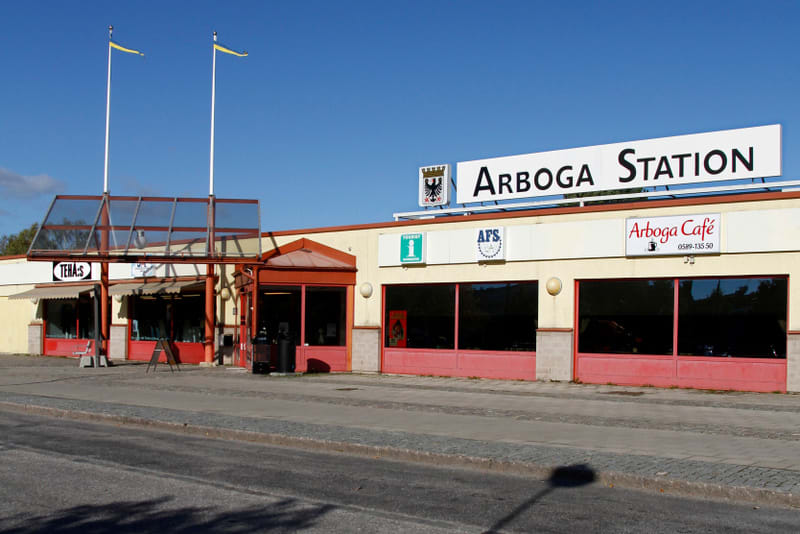 En gul byggnad med röda detaljer och skyltar för "ARBOGA STATION", "Arboga Café" och "TEHA:S". Två flaggstänger med gula flaggor syns mot en klarblå himmel. Framför byggnaden finns en trottoar och en väg.