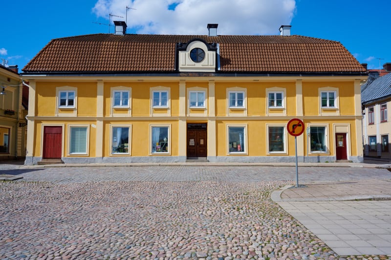 En gul byggnad med brunt tegeltak och vita fönster är centrerad i bilden, ovanför ett torg belagt med kullersten. På en skylt framför byggnaden finns ett förbudsmärke som indikerar "förbud mot att svänga vänster". Blå himmel med moln syns ovanför tak