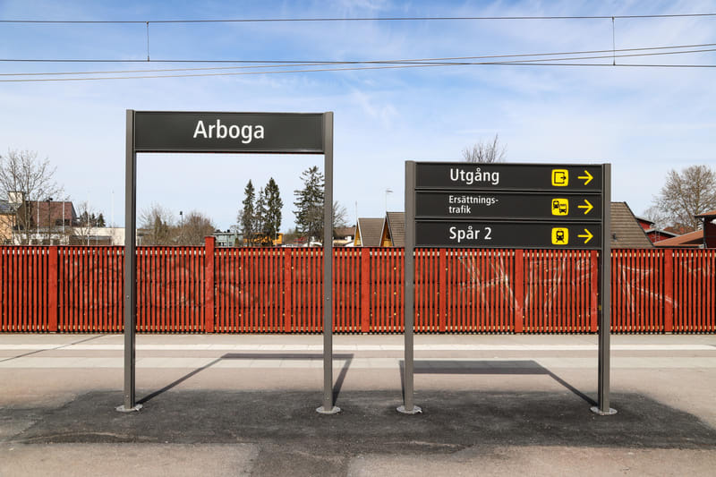 En bild av två skyltar på en tågplattform under en blå himmel. Den vänstra skylten visar namnet 'Arboga' och den högra indikerar 'Utgång', 'Ersättningstrafik' och 'Spår 2' med pilar och symboler. I bakgrunden syns ett rött trästaket, hustak och träd.