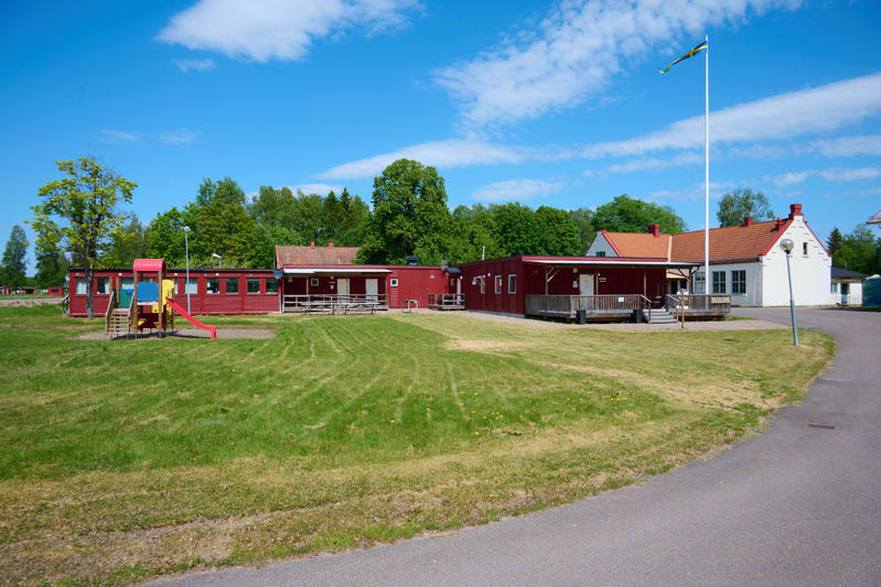En utomhusbild som visar byggnader, gräsmattor och träd under en solig dag. Till vänster syns flera röda barackliknande byggnader och en lekplats med en röd rutschkana. Till höger finns en vit byggnad med rött tak och en flaggstång med en svensk flag