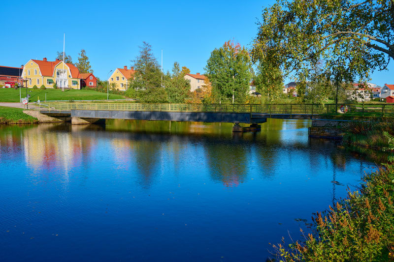 En bildvy över en vattenyta som reflekterar färggranna byggnader och träd. En bro sträcker sig över vattnet, och på horisonten syns bostadshus med gräsmatta framför. Klart blå himmel syns ovanför.