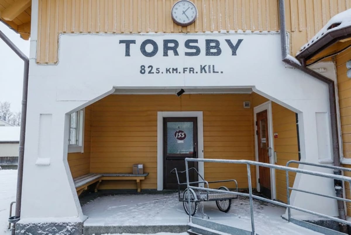 En gul byggnad, troligen en station, med snö på marken. Fasaden har texten "TORSBY 825. KM. FR. KIL." samt en klocka. En dörr med en skylt märkt "ISS" syns, och till vänster om dörren finns en bänk med ett paket. En handkärra står framför dörren.