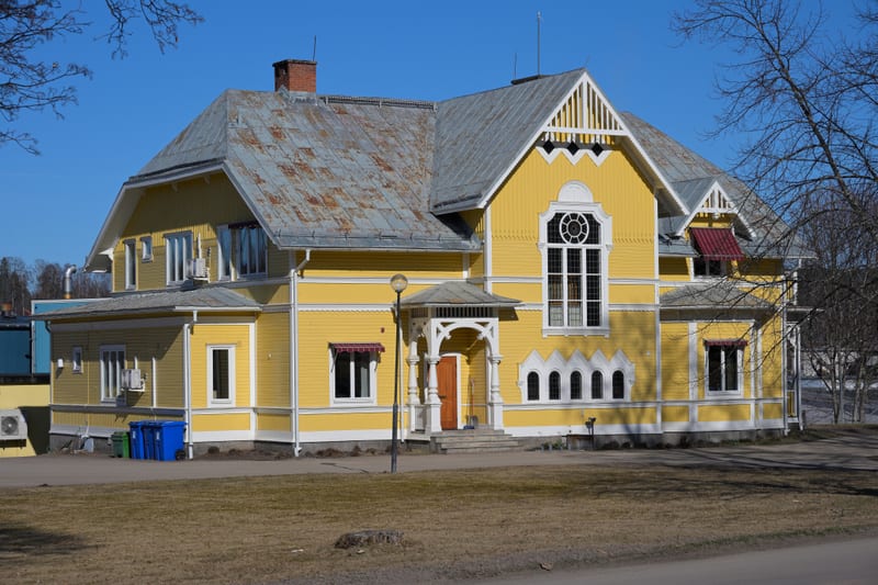 En stor, gul byggnad med vita snickerier och ett plåttak med tegelpipa under klarblå himmel. Framför byggnaden syns torrt gräs, en stubbe och tre soptunnor, samt några bara träd i bakgrunden.