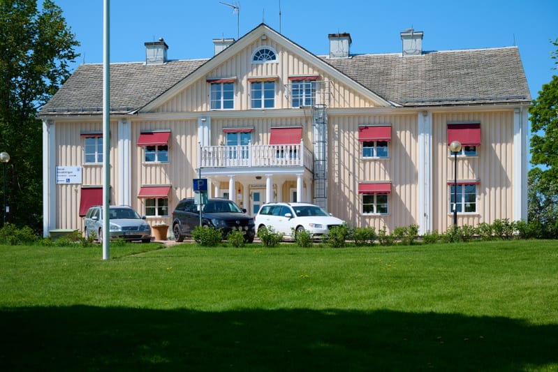 En gul trähusbyggnad med vit trim och röda markiser syns under en blå himmel. Framför byggnaden finns en gräsmatta, buskar och tre personbilar parkerade. En flaggstång står till vänster om byggnaden.