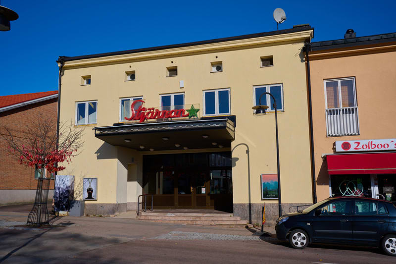 En gul byggnad med skylten "Stjärnan" vid ingången, en satellitparabol på taket. Bredvid ligger en orange byggnad med skylten "Zolboo's". En mörk bil står parkerad framför byggnaderna. En rödbladigt träd och en gatlykta är synliga. Blå himmel är i ba