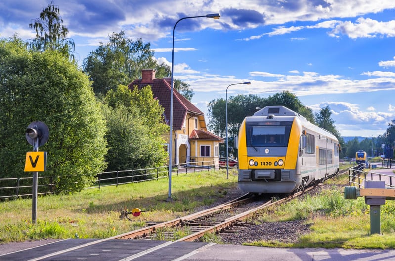 En gul och grå tågvagn syns på en järnväg med en gul byggnad med rött tak i bakgrunden. Ett tågstoppskylt med ett 'V' syns till vänster. Himlen är blå med vita moln och marken är grön med växter. Det finns också träd och stolpar utspridda i bilden.