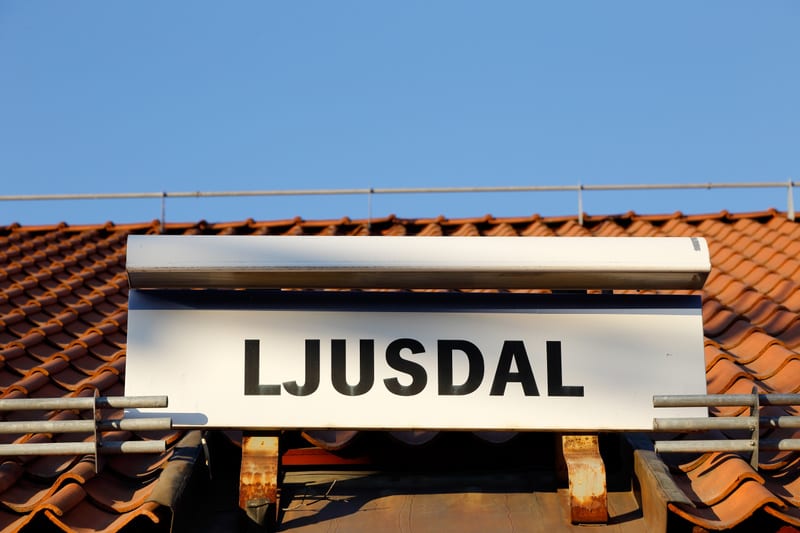 En bild som visar en vit skylt med svarta versaler, där texten "LJUSDAL" är synlig. Skylten är fäst ovanför ett tegeltak med rödbruna takpannor. En blå himmel syns i bakgrunden och delvis en metallräcke ovanför taket.