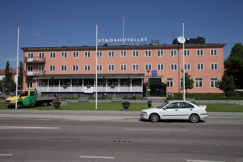 En stor, persikofärgad byggnad kallad "STADSHOTELLET" under en klarblå himmel. Framför byggnaden finns ett grönområde med träd, en parkeringsplats med en grön/gul lastbil och en vit personbil samt en väg i förgrunden.