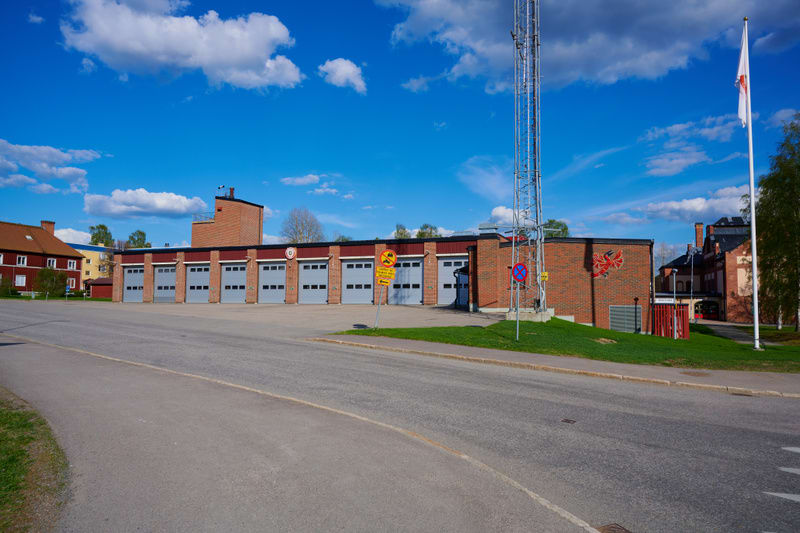 En fasadvy av en brandstation med flera garageportar under en blå himmel med vita moln. Byggnaden är i rött tegel och har röda och grå detaljer. En mast och en flaggstång med en vit flagga syns till höger. Förgrunden består av en asfalterad yta och g