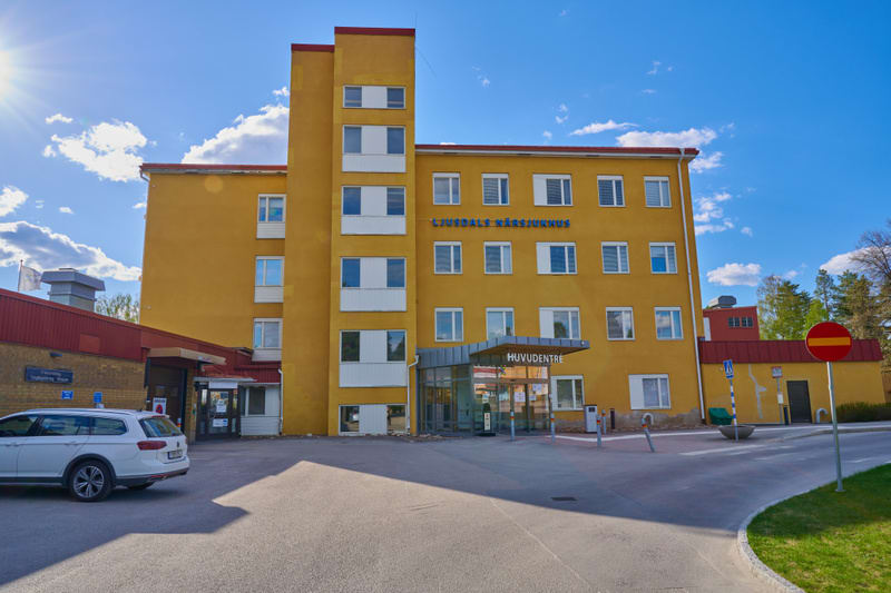 En gul byggnad med många fönster, skyltad som "Ljusdals Närsjukhus" och "Huvudentré", står under en blå himmel med vita moln och solsken. Till vänster syns en vit personbil på en asfalterad yta. Delar av byggnaden är rödbruna och omgivningen har grön