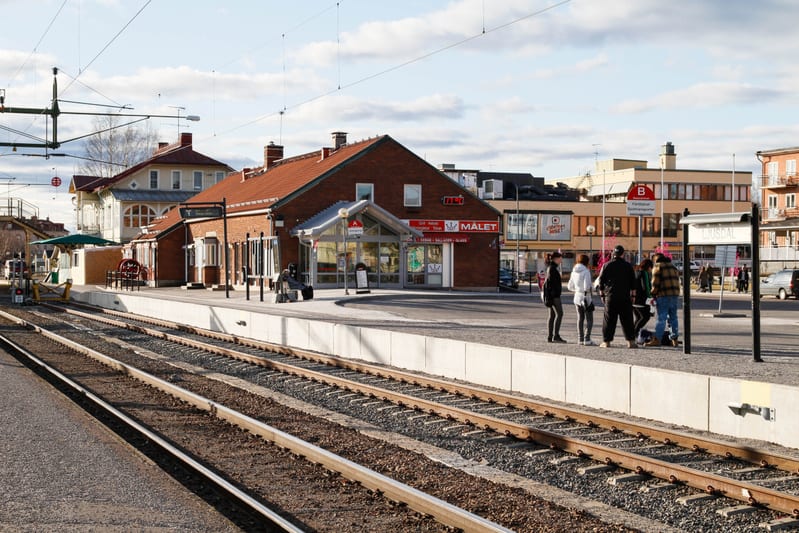 En bild som visar en tågstation med spår i förgrunden och en plattform där flera personer väntar. I bakgrunden syns en tegelbyggnad med texten "MALET" och andra byggnader i en stadsmiljö.