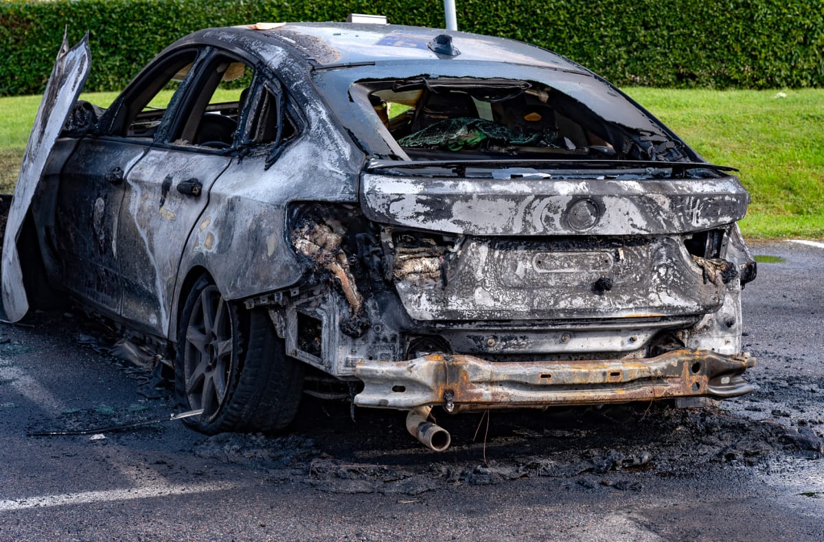 En utbrunnen bil av sedanmodell står parkerad utomhus på asfalt. Bilen är svårt brandskadad med saknade rutor, smält plast och synliga metallkomponenter. Delar av bilen är täckta av sot och aska. I bakgrunden syns grön gräsmatta och en häck.