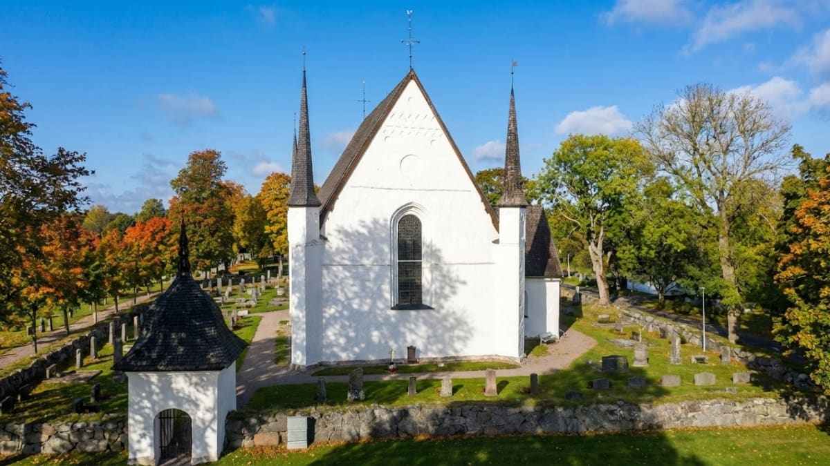 Tierps kyrka | visar en vit kyrka med en kyrkogård runtom, under en klarblå himmel med spridda moln. Träd med löv i höstfärger omger platsen. På kyrkans tak finns små spetsiga torn samt kors- och väderflöjelmotiv.