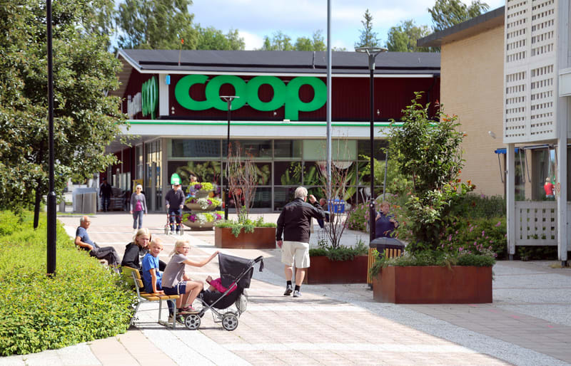 En utomhusbild som visar en Coopbutik och en gångväg framför den. Flera personer, inklusive vuxna, barn och en person med barnvagn, syns på gångvägen. Grönska, blomsterarrangemang och bänkar finns längs gångvägen. Butikens gavel är röd med gröna deta