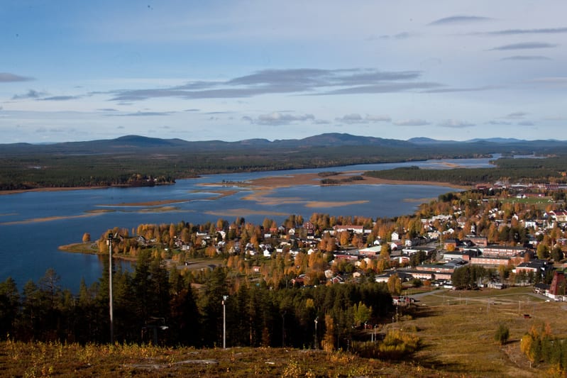 En vidvinkelbild tagen från en höjd visar en klarblå sjö omgiven av barrskog. Till höger om sjön ligger en tätbebyggd ort med varierande byggnader och lövträd i höstfärger. Himlen är blå med utspridda vita moln. I förgrunden syns bergssluttningar.