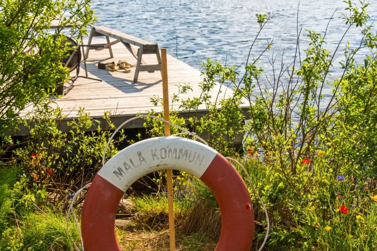 En livboj med texten "MALÅ KOMMUN" hänger på en träpåle i förgrunden, omgiven av grönskande buskar och gräs med några enstaka blommor. I bakgrunden syns en brygga av trä med en bänk och skor, intill en vattenyta.
