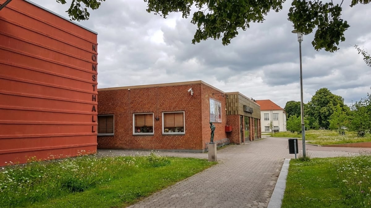 En utomhusbild som visar byggnader med tegel- och putsfasader under en molnig himmel. En av byggnaderna har texten "KONSTHALL" vertikalt på en röd vägg. Förgrunden består av gräs och en gångväg, med träd i bakgrunden.
